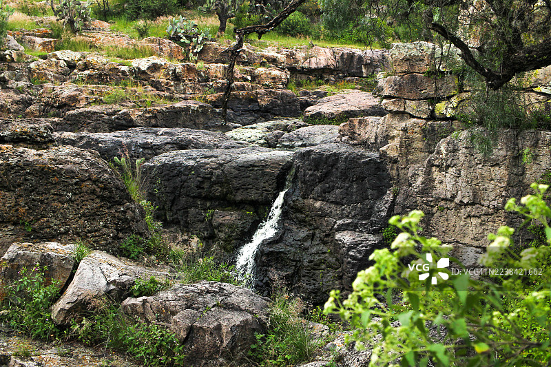 墨西哥圣米格尔-德阿连德的自然景观中，瀑布从深色岩石上倾泻而下，周围绿意盎然。图片素材