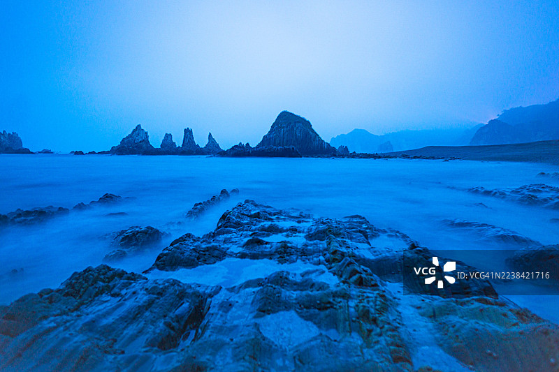 暮色下，西班牙库迪列罗崎岖的海岸线上，海蚀柱和迷雾笼罩的蓝色海洋，以戏剧性的长曝光拍摄。图片素材