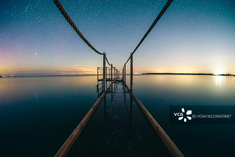 星空下，木质码头延伸入平静的湖面，远方有岸边的灯火，长曝光拍摄图片素材