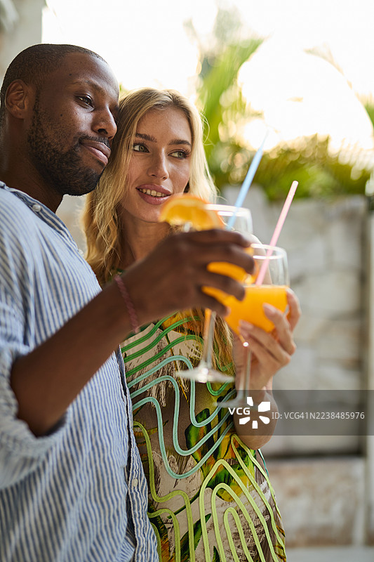 两位朋友在棕榈园里，在阳光下举杯，饮用橙汁，杯中有吸管。图片素材
