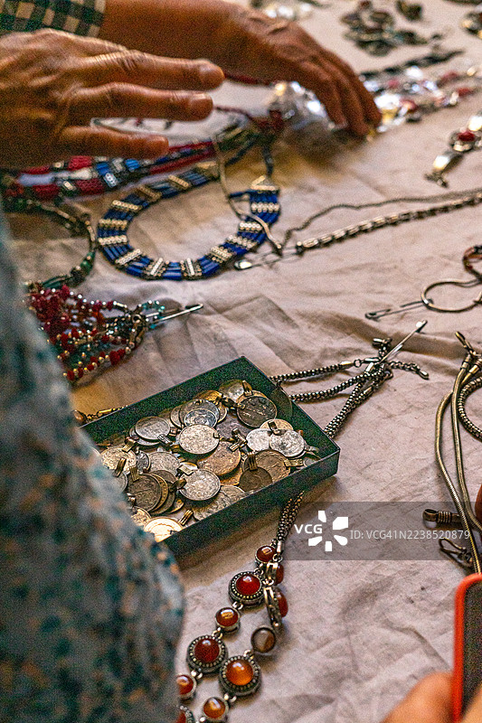 集市摊位上，人们的手正在挑选展示在布料上的手工民族饰品和复古钱币吊坠，聚焦于独特的工艺品和文化艺术品。图片素材
