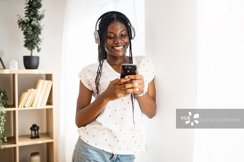 一位戴着耳机、面带微笑的年轻黑人女性，在阳光明媚的房间里，于明亮的起居空间内发短信。图片素材