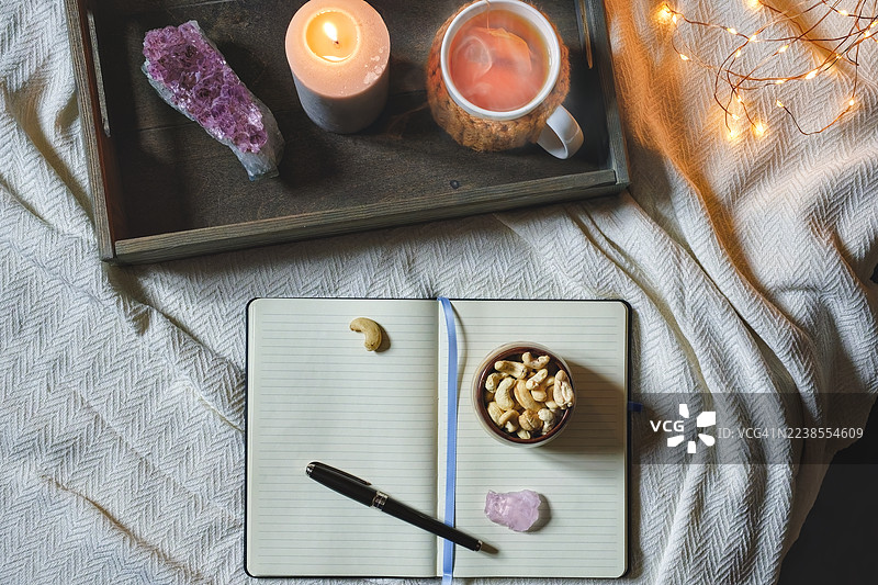 舒适的夜晚，床上放着日记本，配有紫水晶、茶杯、蜡烛和一碗腰果。图片素材