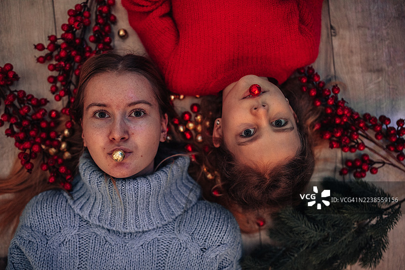 两个孩子躺在地板上，周围散落着红色的浆果、糖果和圣诞装饰品，一位年轻的女性只穿着一件蓝色毛衣，还有一个孩子在舒适的节日氛围中嘴里叼着节日装饰品。图片素材