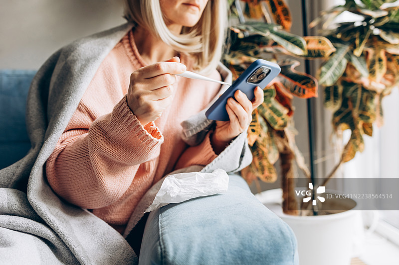 生病的女人在家，裹着毯子测量体温，并用智能手机进行医疗咨询。在线医生咨询、远程医疗服务、流感和感冒症状，以及现代医疗支持的概念。图片素材