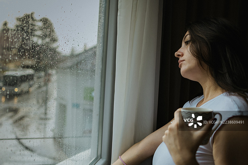雨天忧郁。一位神经多样性、抑郁的年轻女子站在窗边，望着湿漉漉的雨街，手中拿着一杯咖啡。图片素材