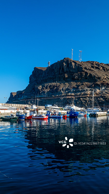 阳光明媚的海港里，渔船停泊在岩石悬崖脚下图片素材