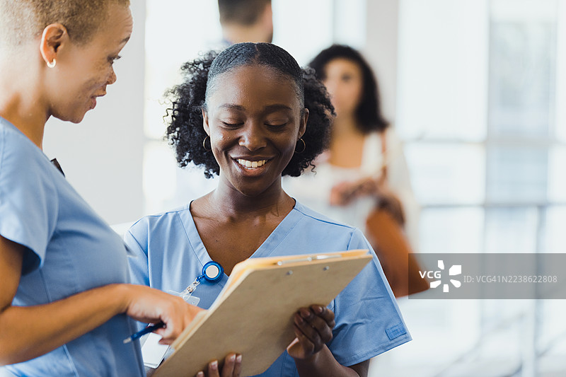 两位穿着浅蓝色手术服的护士在室内微笑着查看剪贴板。图片素材