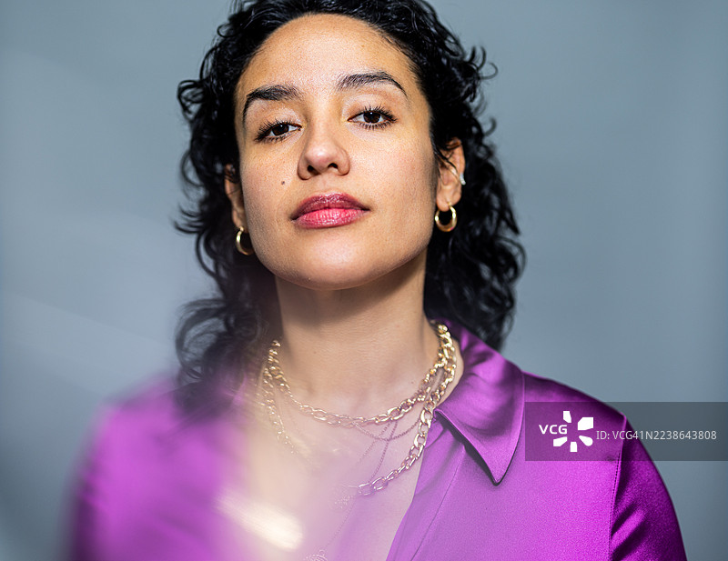 身穿紫色衬衫的年轻西班牙裔女性自信地在灰色影楼背景前摆姿势图片素材