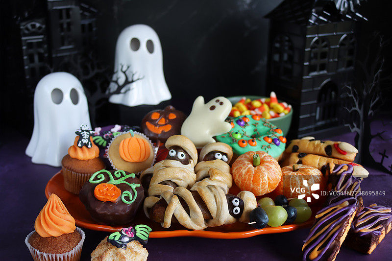万圣节主题派对食物的特写图片，橙色南瓜盘里装着香肠木乃伊（裹着培根的香肠）、橘子南瓜、带眼睛的绿色皇家糖霜甜甜圈、带杏仁指甲的海绵手指饼干，焦点在前景。图片素材