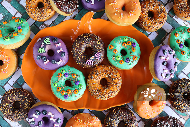 特写照片：万圣节主题的冰糖甜点，装饰着橙色南瓜设计的盘子，有棕色、紫色、橙色和绿色的皇家糖霜甜甜圈，上面装饰着活动眼球、骨头、骷髅和蜘蛛，背景是砖墙效果，俯视角度。图片素材