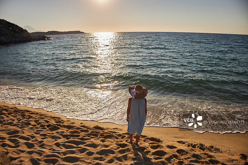 身穿白裙的女子在宁静的海滩上欣赏日落，海浪平静，沙滩呈金色。图片素材