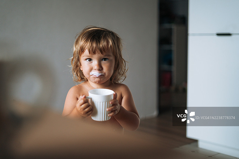 厨房里一个咧嘴笑的有趣小男孩，脸上带着牛奶胡子，这是个在家喝饮料的快乐幼儿。图片素材