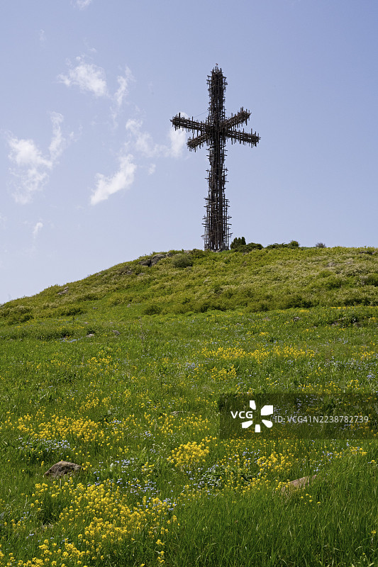 山丘上的大型金属十字架，背景是绿色的草地和黄色的花朵，蓝天下的圣十字纪念碑，由许多小金属十字架组成，亚美尼亚阿拉加措特恩省阿帕兰图片素材