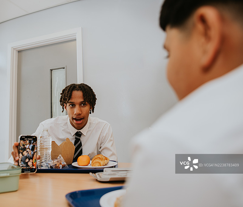 午餐时间，一名高中男生向坐在桌子对面的同学展示一段热门视频。图片素材