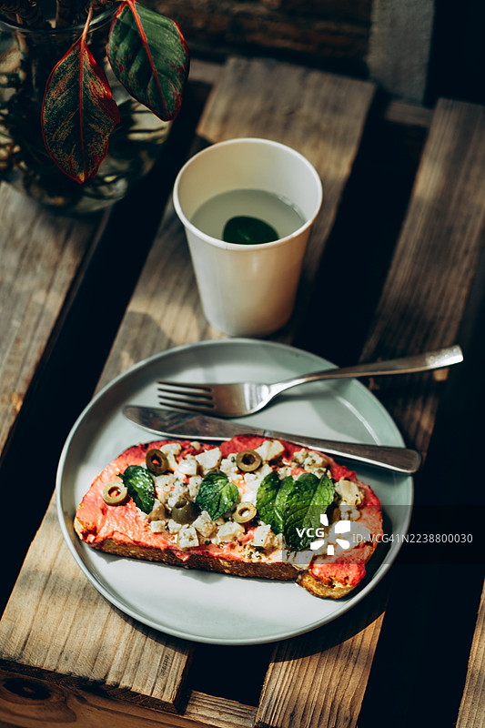 烤面包片上放有甜菜酱、羊奶酪、橄榄和薄荷叶。餐具放在盘子上。摆放在木质表面上。概念：健康的零食，植物性饮食。图片素材