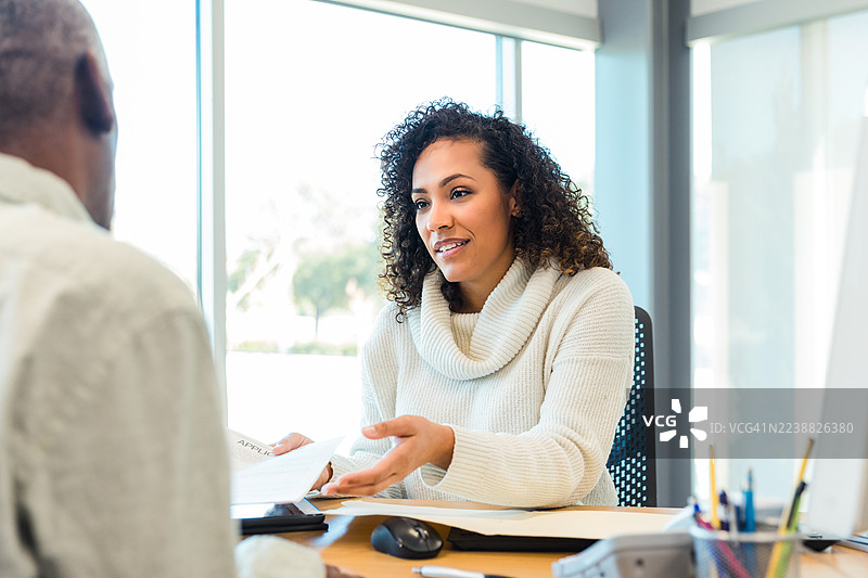 中年女性在室内与不明身份的人交谈，并讨论文件。图片素材