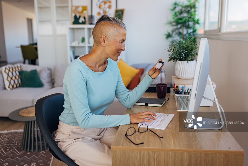 一位女士坐在家里的书桌前，一边使用电脑一边拿着信用卡，享受着在线购物的便利，拥抱数字化生活方式图片素材