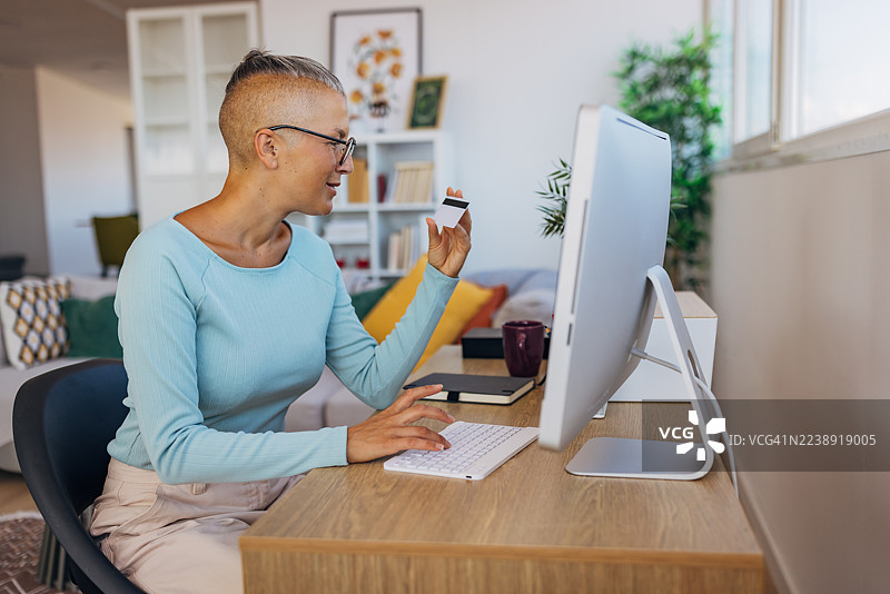 一位女士坐在家里的书桌前，使用台式电脑，手中拿着信用卡。她正在进行在线支付或购物，代表着现代数字生活方式和个人财务管理。图片素材