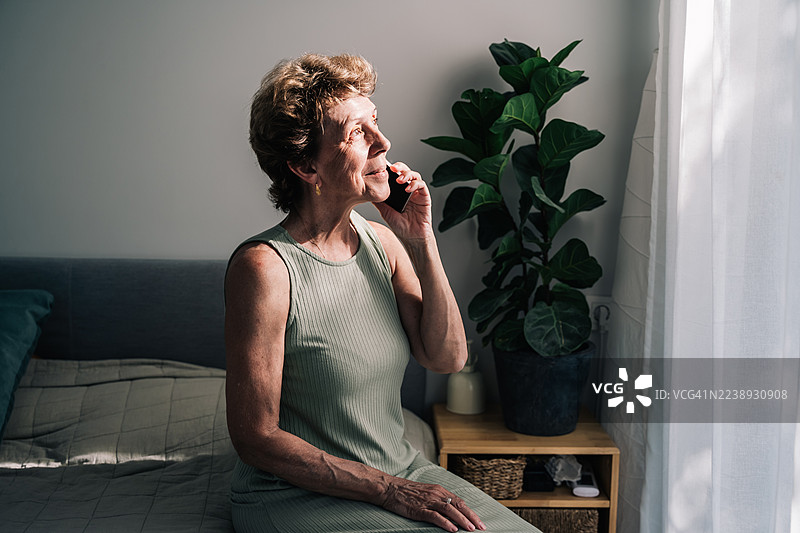 一位年长的女性坐在床边靠窗的位置，正在打电话。房间里洒满了自然光，装饰着柔软的纺织品和绿植。图片素材