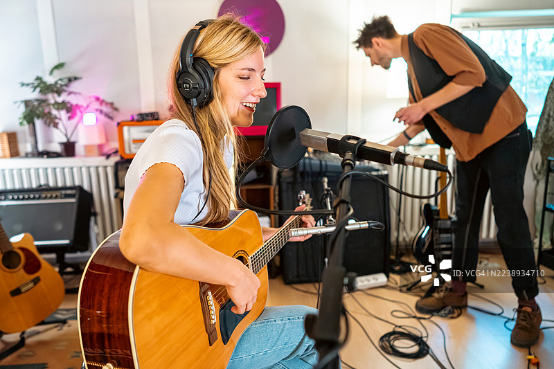 女音乐家在录音棚进行录音，在试音时一边弹奏木吉他一边唱歌，录音工程师在一旁专注地工作。图片素材