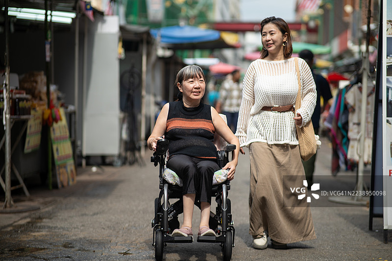 一位亚洲中国女性和她坐轮椅的残疾朋友在城市街道上并肩而行。图片素材