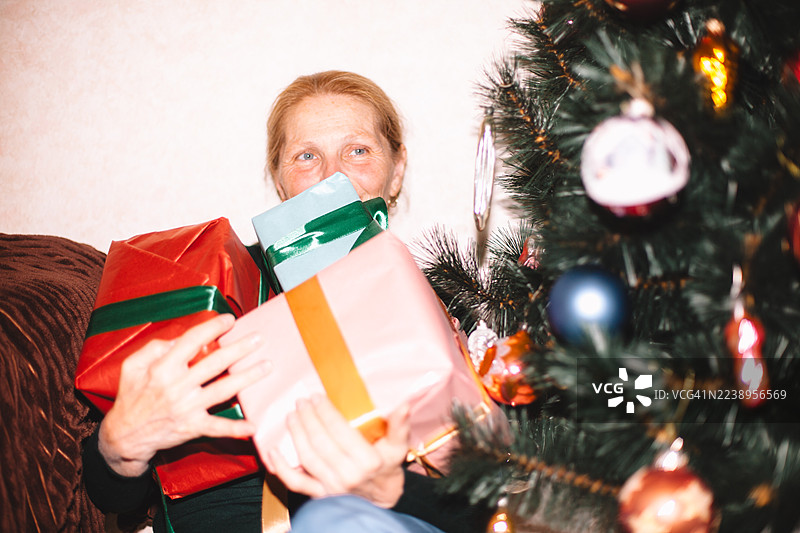 快乐的老年女性在家中圣诞树旁坐着，手里拿着圣诞礼物图片素材