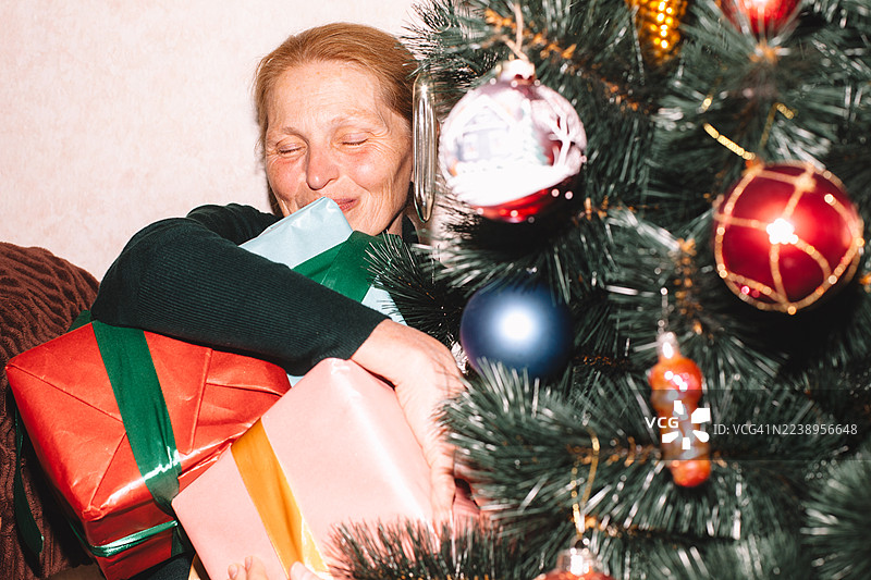 快乐的年长女性在家里圣诞树旁坐着，手中拿着圣诞礼物图片素材