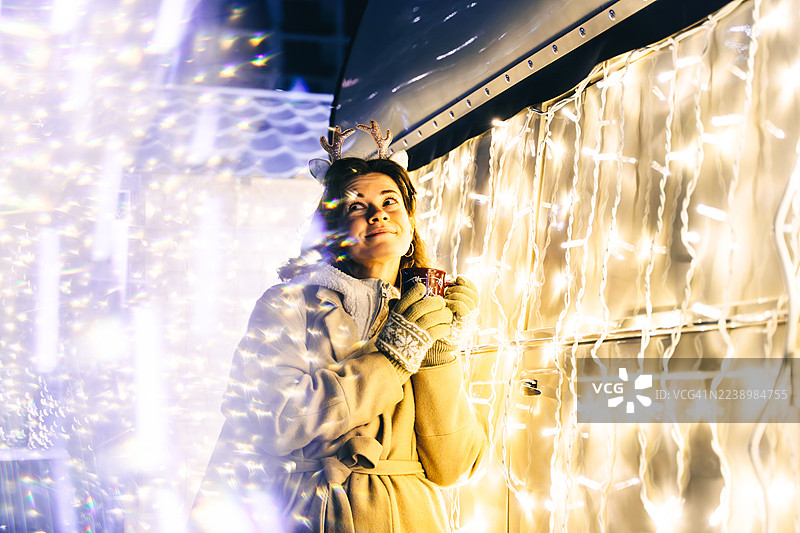 一位女士身穿暖和的外套，头戴驯鹿角，手持一杯热红酒，在夜晚闪耀的节日灯光和散景背景前，拍摄的节日肖像。图片素材