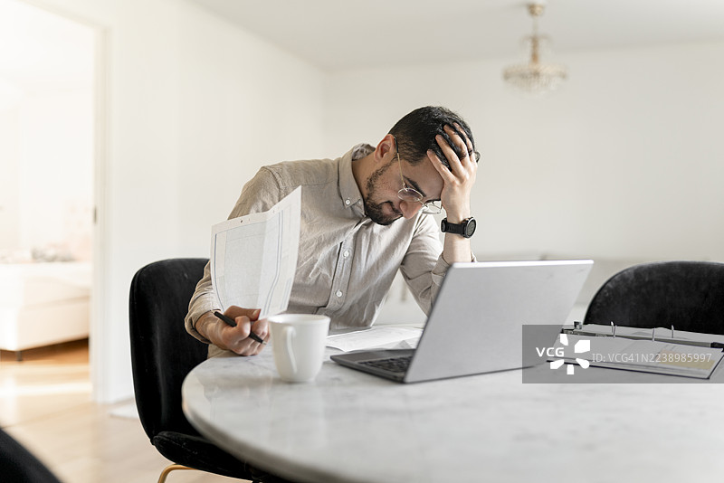 压力山大的男人在家查阅财务报表，笔记本电脑打开，手中拿着文件图片素材