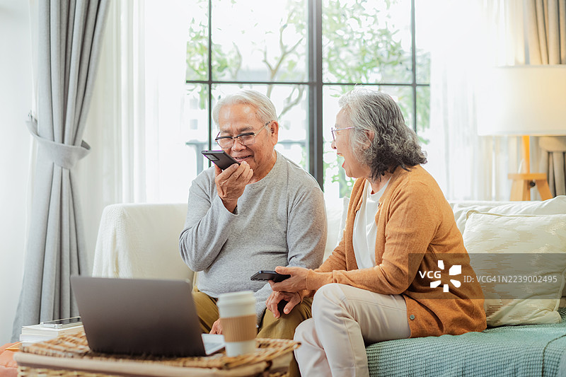 年长的已婚夫妇在家中的沙发上，使用智能手机学习和练习人工智能语音助手及聊天机器人代理，共同提问，怀着喜悦和好奇心探索日常科技学习。图片素材