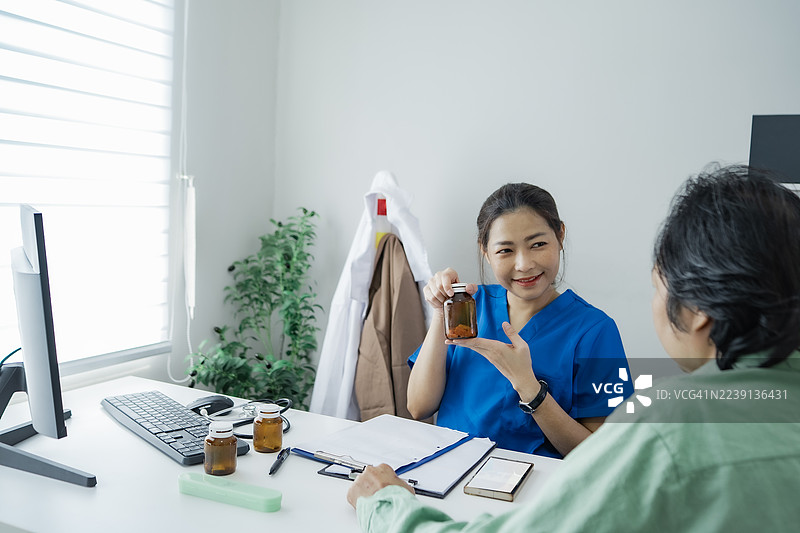 一位自信的女护士穿着蓝色制服，微笑着指向药瓶，在现代诊所办公室里向病人提供医疗建议。图片素材