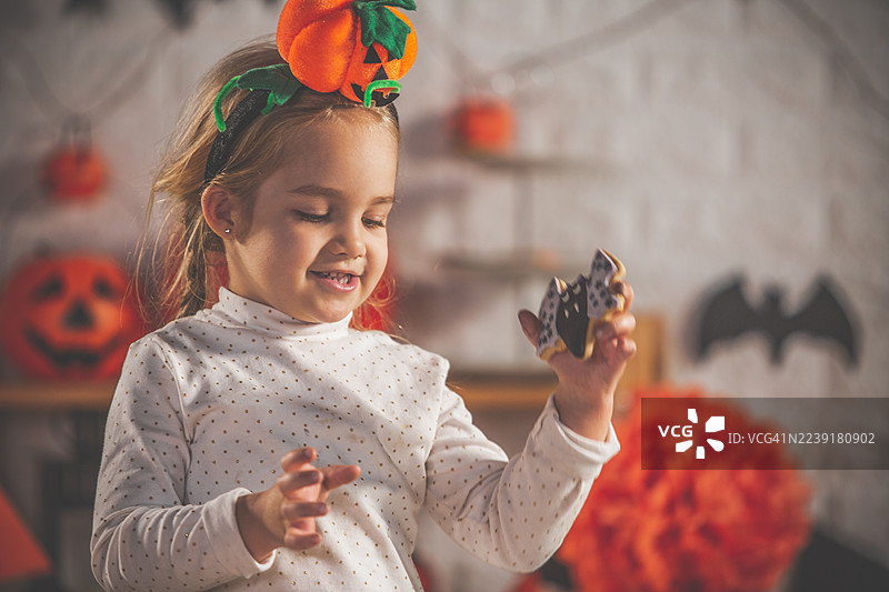 戴着南瓜发带的小女孩，在节日气氛的厨房里，手里拿着万圣节饼干，脸上带着笑容。图片素材