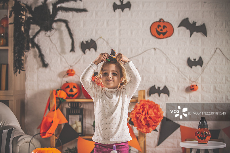 小女孩在家庆祝万圣节，周围有南瓜装饰和蝙蝠旗帜，她微笑着玩着巫师帽。图片素材