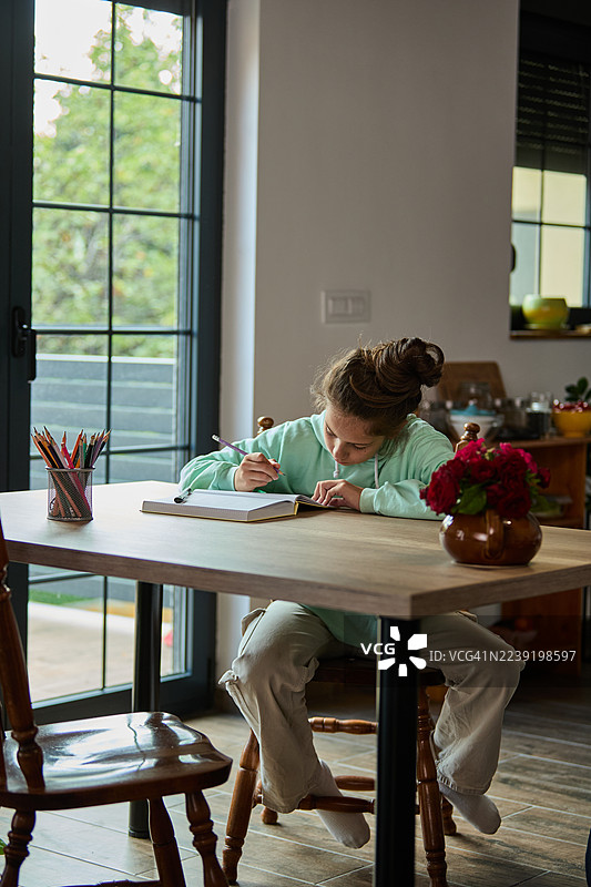 年轻女孩在家木桌旁学习，写着笔记本，旁边放着铅笔图片素材