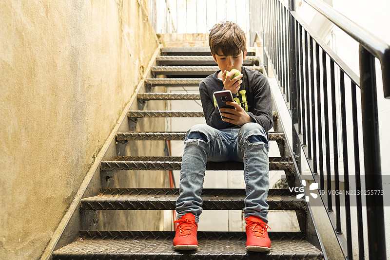男孩坐在城市楼梯上，一边玩手机一边吃苹果。图片素材