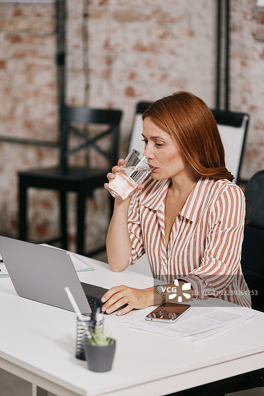 一位女士在现代办公室的白色桌子前工作，一边使用笔记本电脑一边喝水。图片素材