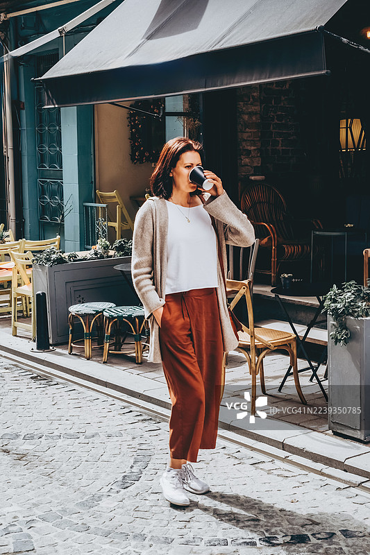 一位穿着休闲服饰的白人女性，在鹅卵石街道旁走过一家咖啡馆，同时喝着手中的黑色杯子。光线明亮，色彩温暖。图片素材