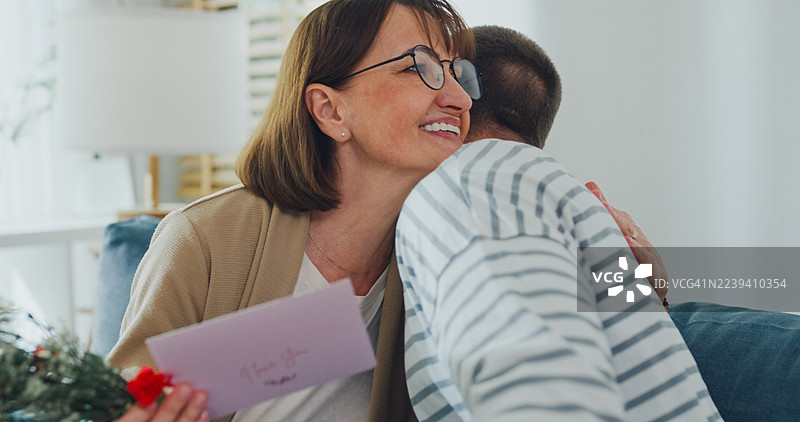 十几岁的白人儿子在客厅沙发上送花给惊喜的妈妈。家庭生活方式和母亲节概念。图片素材
