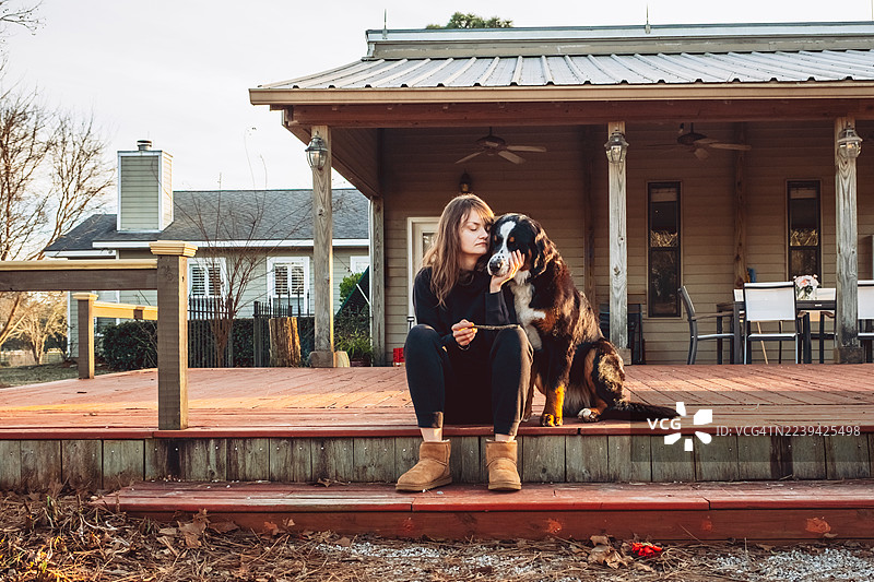 身穿黑色服装、脚踩棕色靴子的年轻女子坐在门廊台阶上，旁边是一只伯恩山犬。舒适的木屋环境。田园生活舒适感、宠物、家庭生活概念。图片素材