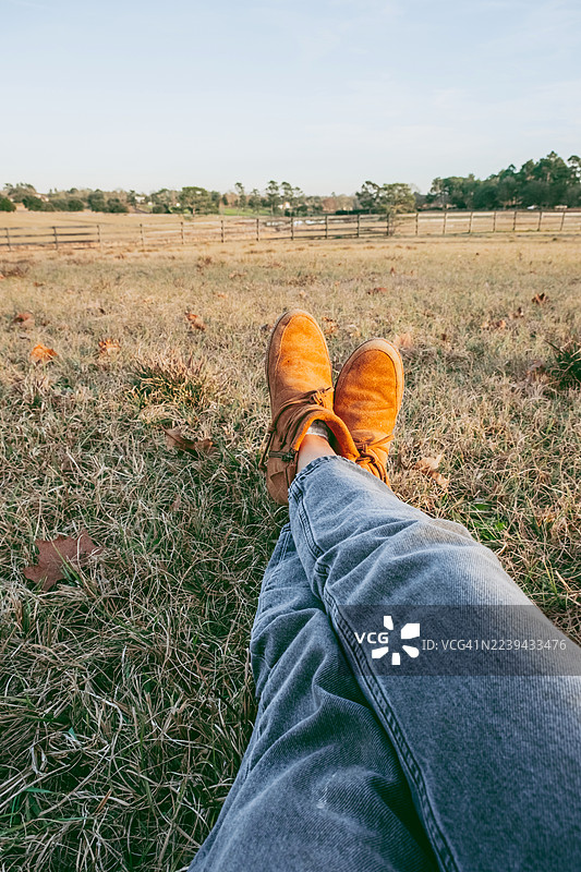 身穿牛仔裤和橙色麂皮靴的人躺在干草地上，双腿交叉。远处是宁静的田野和木栅栏的宽阔视野。乡村的休憩，慢生活，宁静的独处。图片素材