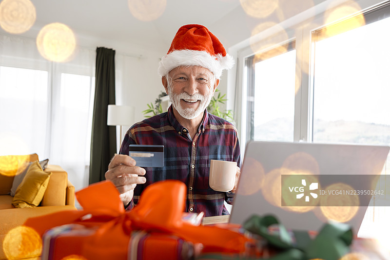 一位戴着圣诞帽的老人在家通过信用卡在网上购买圣诞礼物，他正看着镜头。图片素材