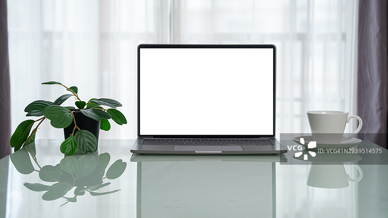 在家里的办公桌上放着一台笔记本电脑。在极简风格的办公室里，一张木制工作桌上放着一台笔记本电脑，屏幕是白色的空白，旁边还有咖啡杯和盆栽植物，以及一些装饰品。图片素材