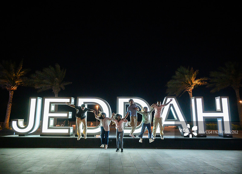 夜幕下，杰达发光标志前，一群朋友在空中跳跃的广角镜头图片素材