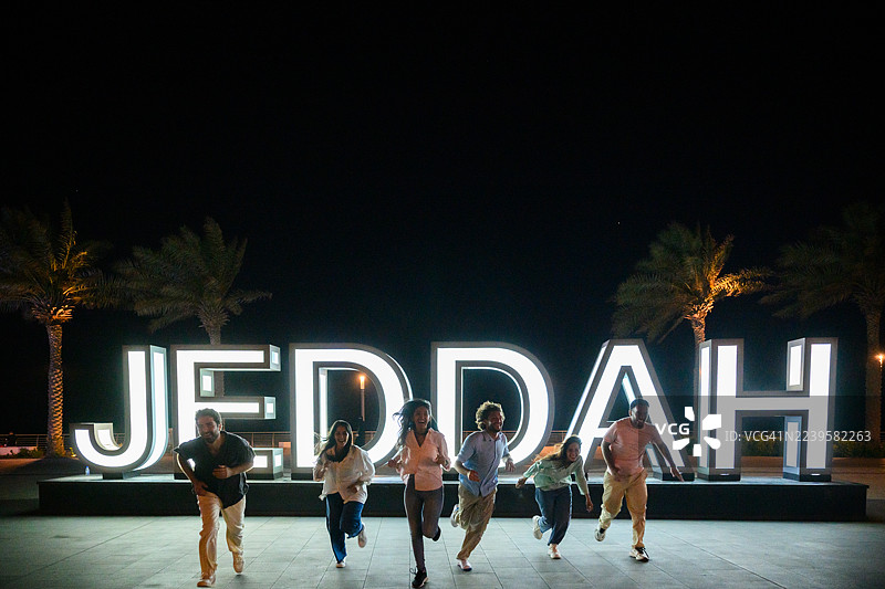 夜间，一群朋友在灯火辉煌的吉达标志旁欢乐奔跑的广角镜头图片素材
