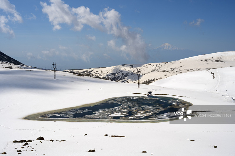 白雪覆盖的山丘环抱着冰封的山间湖泊，天空晴朗，背景是土耳其的亚拉腊山（Ashot Orujyan Lake），以及海拔4090米的已灭绝的平流火山阿拉加茨山（Aragaz, Aragats, Aragac, Alagyaz），位于亚美尼亚的阿拉加措特恩省和希拉克省。图片素材