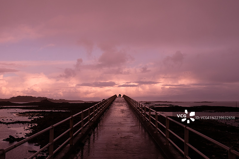 在法国罗斯科夫，夕阳染红天际，粉紫色的晚霞将海面上的长桥笼罩在一片宁静而神秘的氛围中，两人漫步桥上，构成一幅宁静而神秘的画面。图片素材