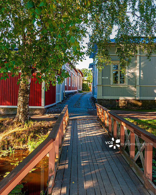 木桥通往鹅卵石小路，两侧是色彩鲜艳的传统房屋，周围环绕着郁郁葱葱的树木，位于芬兰一座历史悠久的欧洲城镇。图片素材