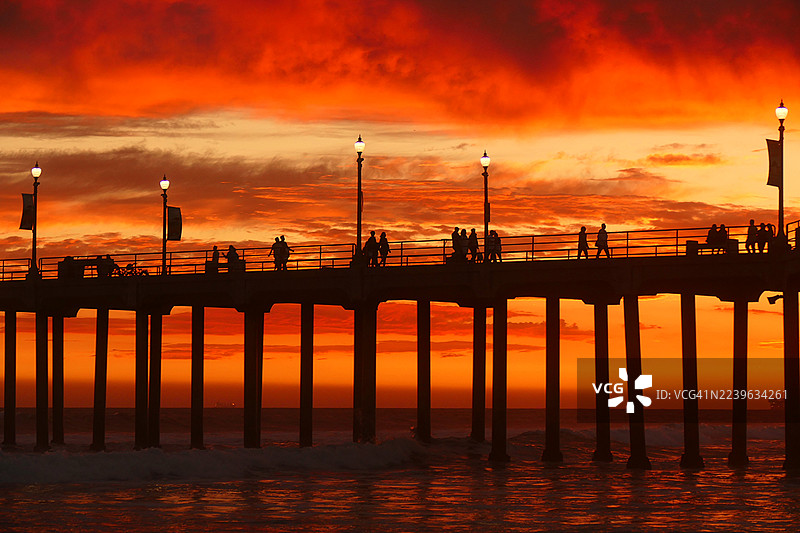 在海边，夕阳将天空染成鲜艳的橙色，码头上的人影剪影，美国亨廷顿海滩图片素材