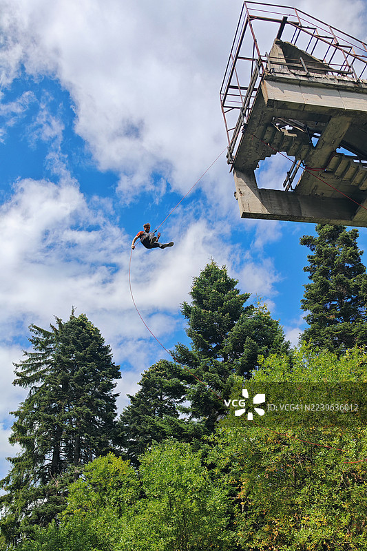 男人在高大建筑旁利用绳索下降，周围有树木和蓝天，这是一项冒险的户外活动。图片素材